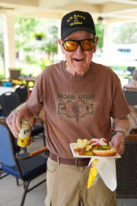 Smiling elderly U.S. Navy veteran wearing sunglasses and a Moab, Utah t-shirt, holding a plate of food and a drink at Concordia on the Lake – Apartment Cottage and Homes